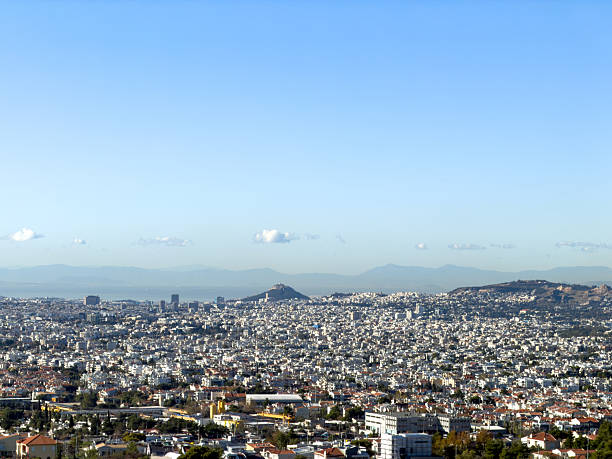 Panoramic Athens cityscape with Mount Lycabettus, Greece. Dense white buildings and distant mountains under blue sky, viewed from a high lookout.