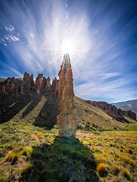 Dramatic scene of sunlight radiating behind tall rock formations, creating long shadows across the valley floor. The glowing sky adds energy and contrast to the rugged Patagonian terrain.