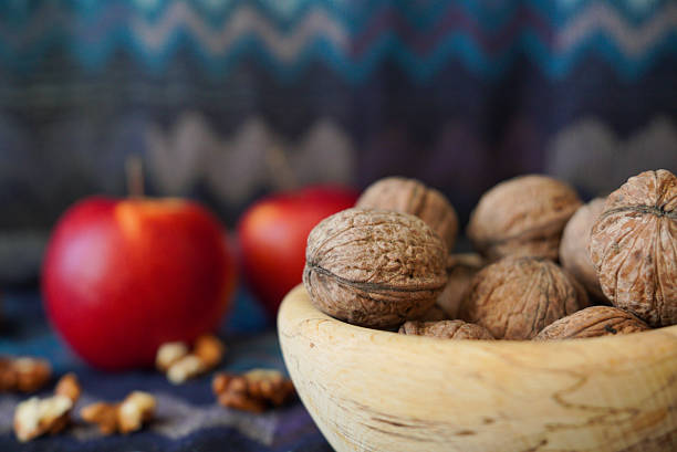 Walnuts and walnut kernels in a wooden bowl with red apples in the background. Rustic still life food photography, healthy nutrition concept, natural organic ingredients.