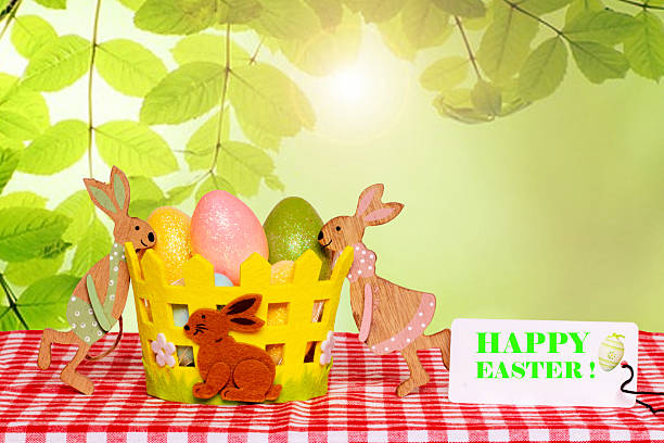 Easter card. Colorful easter eggs in a basket with two wooden bunnies and a happy easter tag on a red checkered tablecloth over abstract spring background. Religion and culture concept