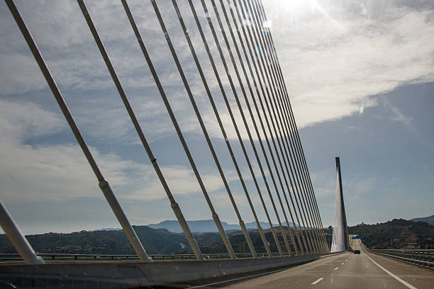 A wide-angle shot of a modern cable-stayed bridge with white steel support beams stretches across an empty road under a clear blue sky, bordered by trees in autumn hues and a calm rural landscape