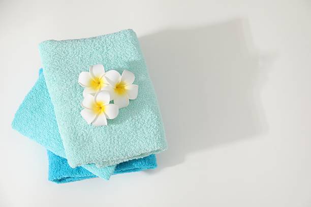 Stack of colorful towels and plumeria flowers on white table, top view. Space for text