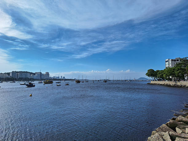 A wide view of a calm coastal bay with small boats anchored across gently rippling water, framed by an urban waterfront in the background. Buildings, shoreline vegetation, and a stone embankment define the city’s relationship with the sea under a clear blue sky with soft clouds. The image conveys tranquility, coastal lifestyle, and everyday maritime activity, making it suitable for concepts related to travel, waterfront living, urban nature, tourism, and coastal cities in Brazil or similar regions.