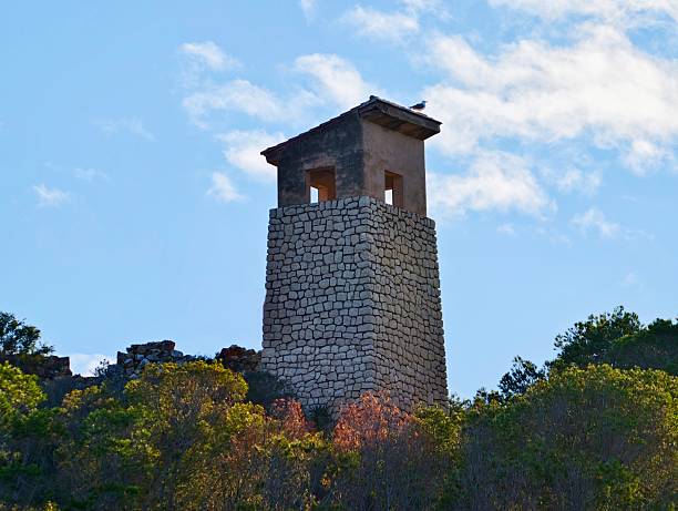 Stone Watchtower Atop Wooded Hill Overlooks Rolling Valley Under Bright Blue Sky With Scattered Clouds.