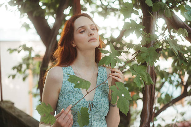 Young woman with red hair standing outdoors, gently touching green grape leaves, feeling serene in natural light