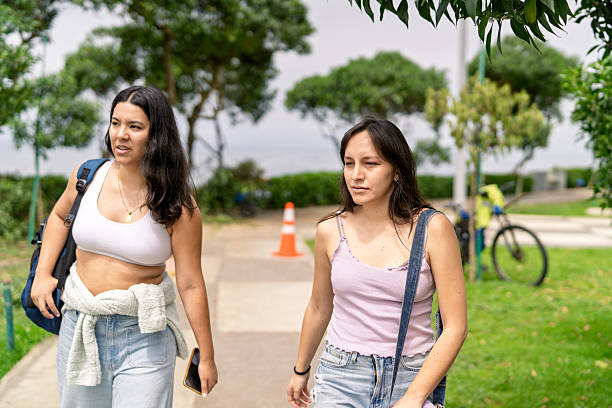 Two young women friends enjoying a relaxing stroll on a paved path in a green park by the sea, unwinding from daily routine