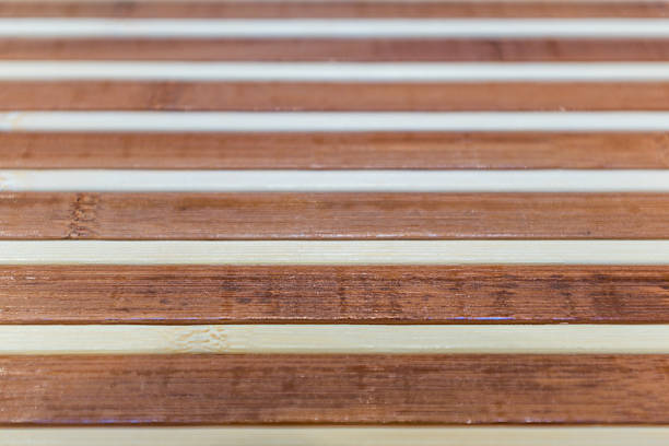 This close-up image focuses on the rhythmic pattern of a bamboo mat. The design features wide, polished slats of dark reddish-brown bamboo alternating with thinner, light beige strips. The natural grain and characteristic nodes of the bamboo are clearly visible on the darker pieces, adding organic texture. The shallow depth of field creates a blur in the foreground and background, isolating the sharp details in the center and highlighting the smooth, varnished finish of the material.