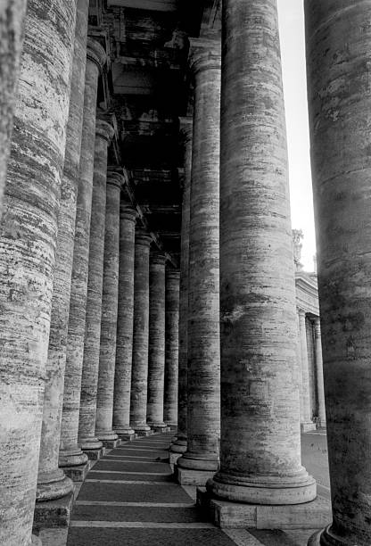 Black‑and‑white photo of a sweeping colonnade formed by evenly spaced cylindrical stone columns supporting a horizontal entablature. The curved alignment and repeating structural rhythm create strong symmetry, while the weathered surfaces and stone‑paved walkway emphasize the age and texture of the architecture.