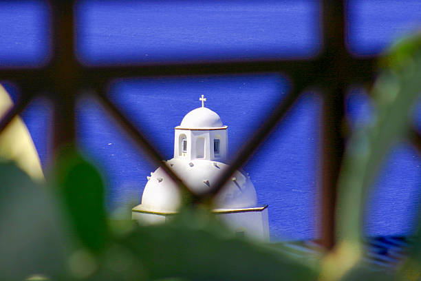 White domed Greek Orthodox church seen through blurred railing with deep blue sea in background, dreamy Mediterranean island scene.