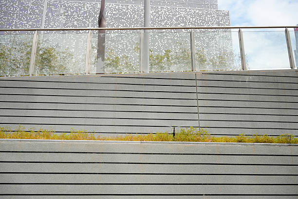 A striking architectural detail of a modern urban building featuring a perforated metal facade, transparent glass railings, and horizontal concrete retaining walls integrated with low-growing greenery. The design combines industrial materials with natural elements, creating a harmonious balance between structure and nature. This minimalist yet functional landscape showcases contemporary trends in sustainable city planning, public space design, and innovative façade systems. Ideal for illustrating themes such as urban renewal, eco-friendly architecture, and modern infrastructure.