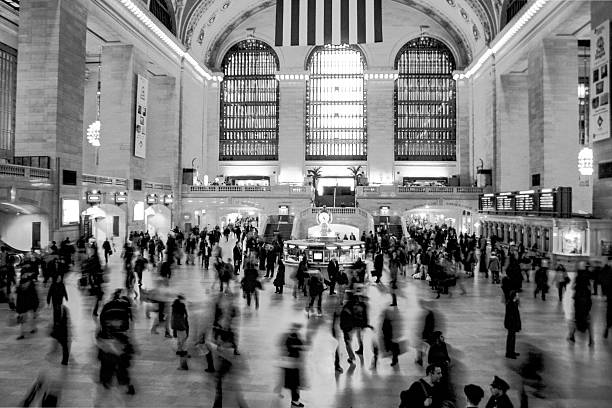 Grand Central Terminal interior in monochrome, crowded concourse with blurred commuters beneath huge arched windows and American flag.