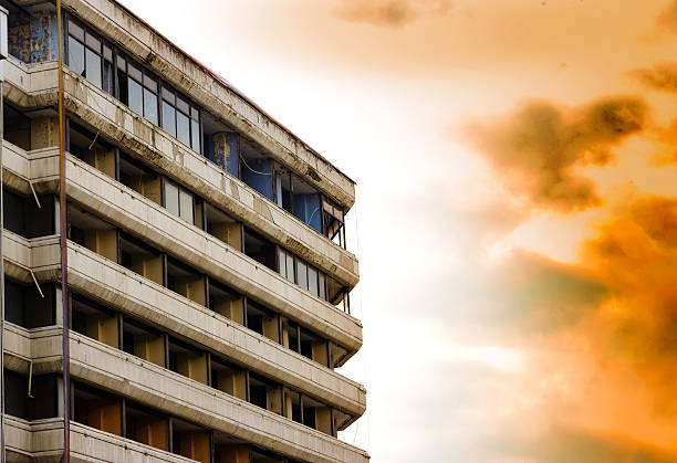 Abandoned multi-story building with peeling facade, broken windows and exposed interiors under dramatic orange and gray cloudy sky, featuring horizontal balconies and urban decay atmosphere.