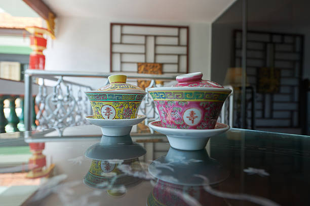 Two vintage Peranakan Nyonya-style porcelain tea cups with lids (gaiwan) featuring colorful traditional Chinese longevity motifs, placed on a reflective glass table in a heritage house interior with soft natural light and bokeh background.