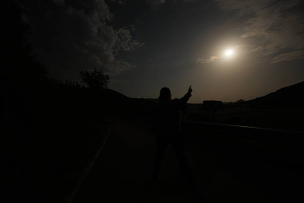 Person's silhouette standing in darkness, pointing gesture towards the bright full moon in the night sky