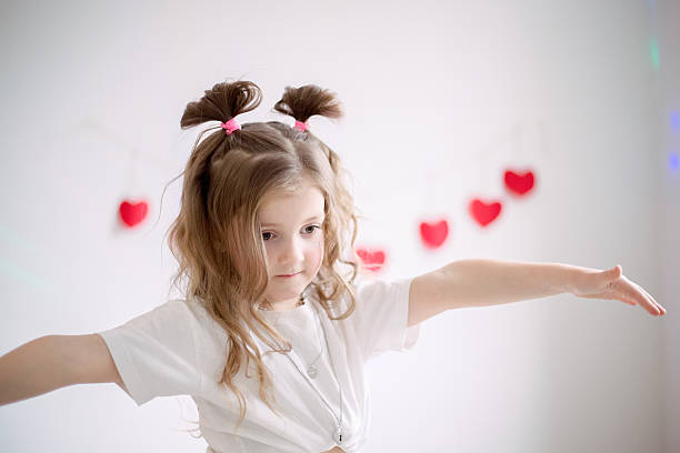 Happy young girl expressing herself through energetic dance movements against white wall decorated with festive red hearts, celebrating Valentine's Day with joy and carefree playful spirit