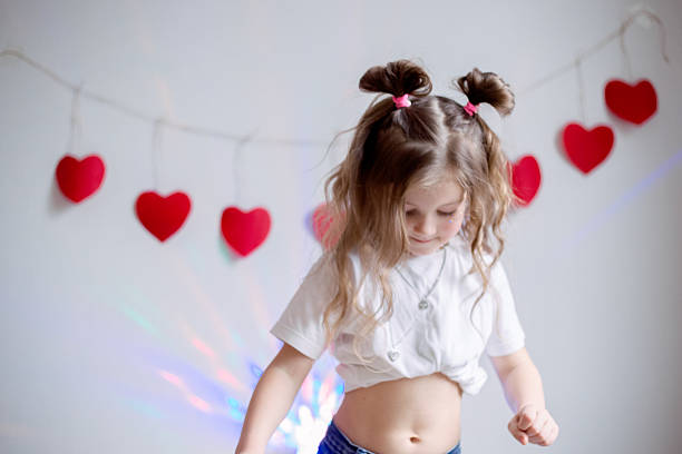 Happy young girl expressing herself through energetic dance movements against white wall decorated with festive red hearts, celebrating Valentine's Day with joy and carefree playful spirit