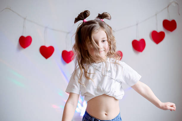 Happy young girl expressing herself through energetic dance movements against white wall decorated with festive red hearts, celebrating Valentine's Day with joy and carefree playful spirit