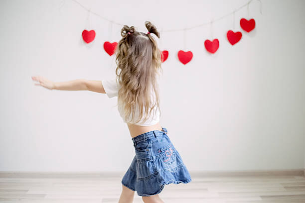 Happy young girl expressing herself through energetic dance movements against white wall decorated with festive red hearts, celebrating Valentine's Day with joy and carefree playful spirit