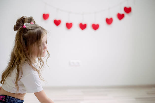 Happy young girl expressing herself through energetic dance movements against white wall decorated with festive red hearts, celebrating Valentine's Day with joy and carefree playful spirit