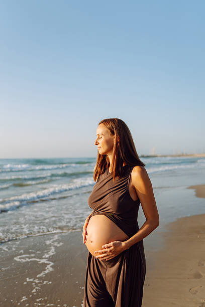 Beautiful woman expecting child stands on beach in sun's rays. Pregnant woman enjoys seascape, feeling freedom of sunset. Concepts of expecting, walking, freedom, and relaxation.