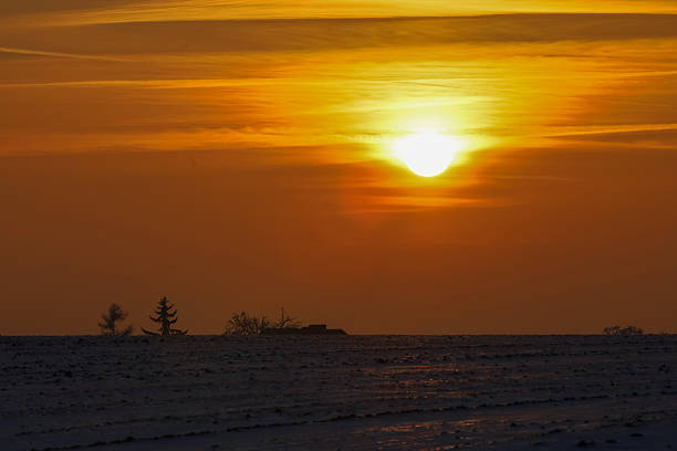 The sun casts a warm, golden glow across the sky, illuminating thin clouds above a frosty landscape with sparse vegetation and a small structure on the horizon