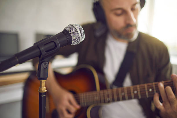 Musician playing guitar and singing into mic. Close up shot in soft, selective focus of modern microphone in recording studio, with man playing some quiet music in blurred background. Music concept