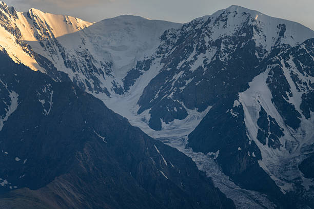 Close view of a mountain glacier flowing between steep snow-covered peaks in the Altai Mountains under soft natural light. Remote alpine landscape with no people, suitable for climate, nature, and environmental themes.