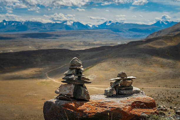 Rocky viewpoint with two stacked stone cairns in the foreground and a wide view of the Kurai Steppe and distant Altai Mountains under a partly cloudy sky. Outdoor landscape with no people, suitable for travel, nature, and background use.