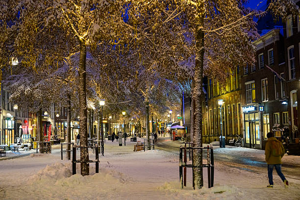 This winter photograph captures the Melkmarkt in Zwolle with a christmas decoratioins in the shopping street in Zwolle. Fresh snow blankets streets. Warm amber lighting contrasting beautifully with the cool blues and whites of the snowy surroundings. Narrow streets radiate outward, glowing with lines of streetlights that guide the eye through the dense urban fabric. Gabled houses add layers of history and texture, while the distant cityscape fades gently into the winter dusk. Overall, the image conveys a sense of quiet grandeur, where centuries-old brickwork meets the stillness of a winter evening in the city.