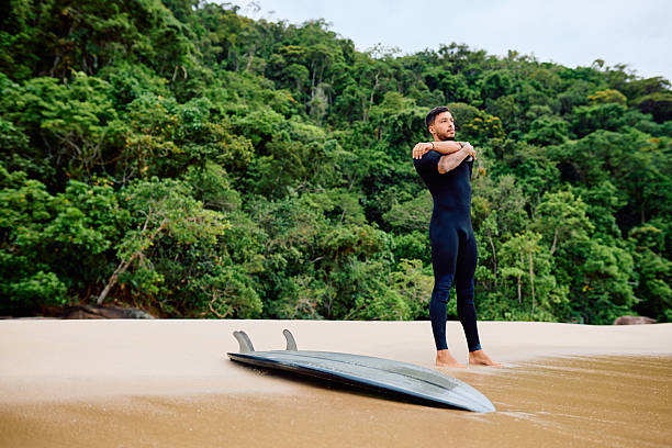 Surfer man in wetsuit stretching arms on a sandy beach, preparing for surfing amidst lush green tropical jungle