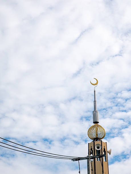 Islamic mosque minaret tower with golden crescent moon symbol and loudspeakers under blue sky and white clouds, religious architecture concept.