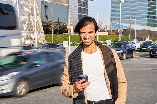 Man standing outdoors in a bustling city, smiling and looking at camera while holding a smartphone with moving cars in background