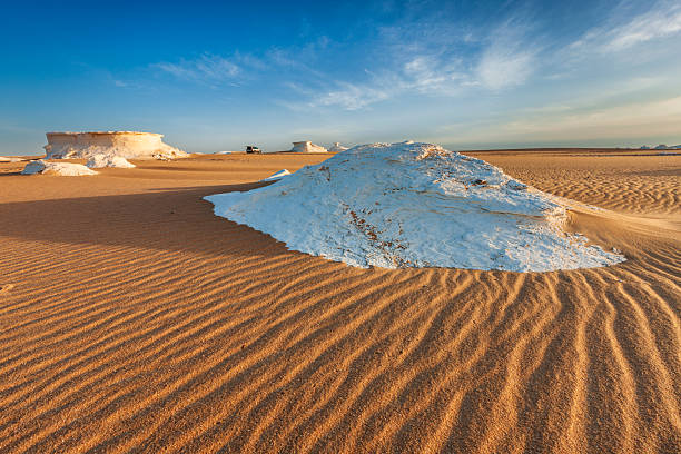 A wide-angle view of a sunlit desert plain dotted with low, white, mushroom-shaped rock formations. Golden rippled sand fills the foreground and leads the eye toward larger sculpted outcrops on the horizon; a small off-road vehicle provides scale. Clear blue sky with wispy clouds and warm late-afternoon light emphasize the textures and contrasts between sand and stone.