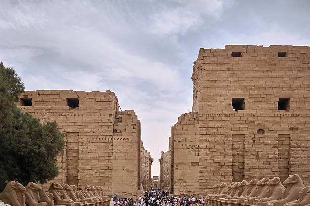 Wide view of monumental sandstone temple complex featuring towering pylons, symmetrical architectural geometry, and a central processional path lined with ram-headed sphinx statues. Luxor, Egypt