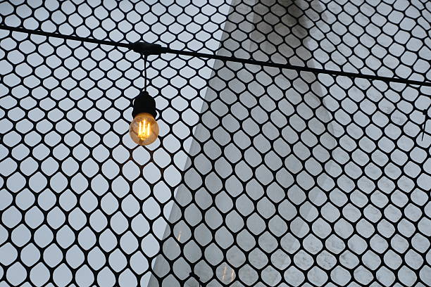 Single warm light bulb hanging in front of wire mesh fence with soft sky background