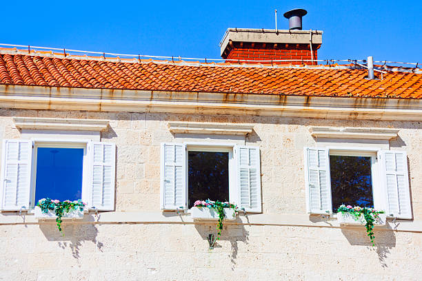 White stone building with tiled roof stands against clear sky. Three windows with white shutters and flower boxes add Mediterranean charm