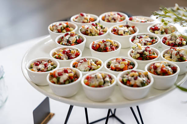 Elegant display of small white bowls filled with colourful vegetable salad, garnished with pomegranate seeds and creamy dressing, presented on white round platter on modern stand, ideal for buffet or corporate event