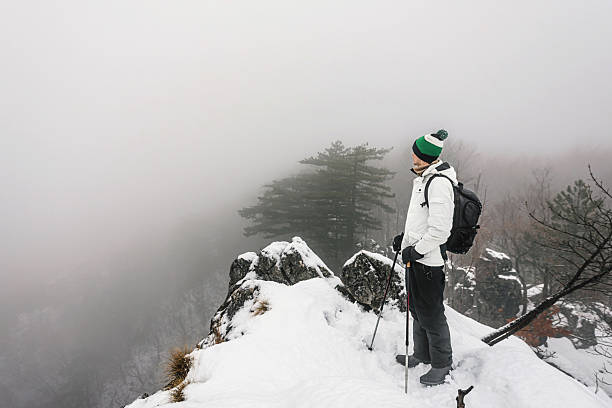 Adventurer in winter hiking gear with trekking poles, hiking on snowy rocky mountain ridge in foggy conditions. Man walking in steep path on rocky outcrop. Winter travel adventure in cold landscape.