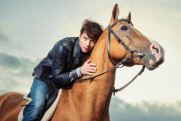 Young man in a leather jacket hugs and caresses a horse outdoors