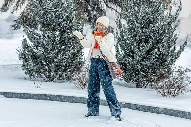 Young woman in warm winter clothing standing in a snowy park and catching falling snow during heavy snowfall. Atmospheric outdoor winter scene symbolizing cold weather, nature, seasonal mood, leisure, and cozy winter emotions.