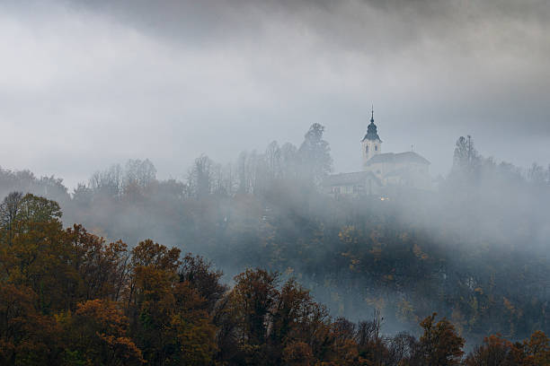 small hilltop church emerging above a dense autumn forest covered in mist, creating a peaceful and atmospheric landscape. warm fall colors, soft fog, and the elevated position of the church highlight the harmony between nature and architecture.