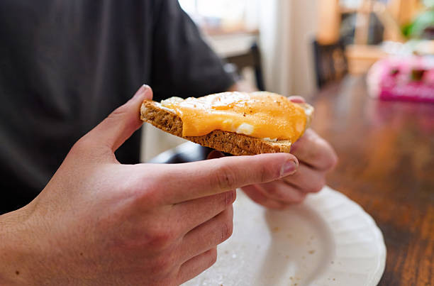 Side view of a young man holding an open-faced fried egg and melted cheddar toast above a plate at a wooden dining table. Cozy, unhurried lunch mood with soft background blur and copy space on the table for text.