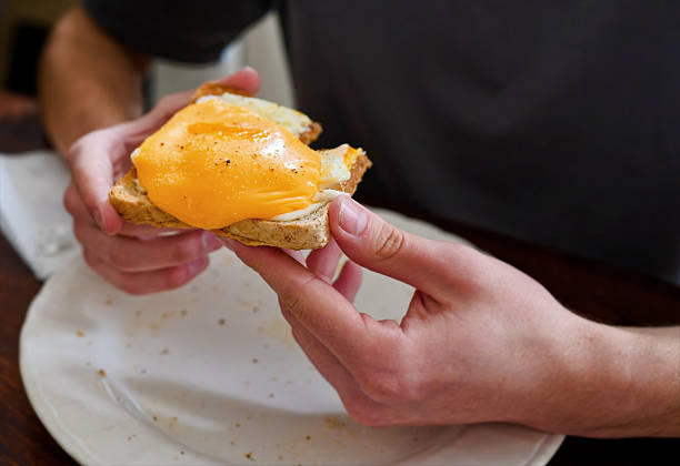 Close-up of hands holding an open-faced sandwich topped with fried egg and melted cheddar over a plate with crumbs, captured in a casual at-home lunch moment. Clean, minimal background feel with copy space for food or lifestyle messaging.