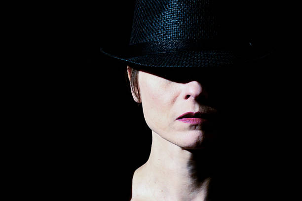 Low-key portrait of a woman partially hidden in shadow, wearing a black hat against a dark background.