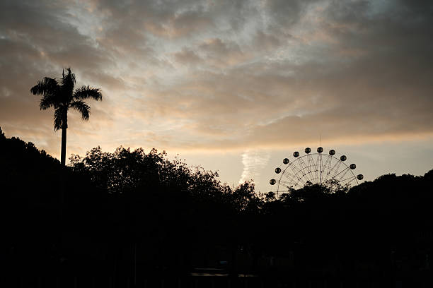 A striking silhouette of an amusement park Ferris wheel stands against a dramatic sunset sky, where bands of deep orange and gold blend with layers of dark storm clouds. The wheel’s defined structure contrasts with the soft gradient of the horizon, framed by distant tree outlines. This low-angle shot emphasizes scale and mood, capturing a moment of serene yet powerful transition between day and night—ideal for themes of nostalgia, urban leisure, and natural spectacle.