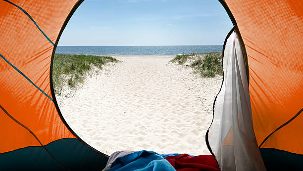 Point of view from inside an orange camping tent looking out onto a bright sandy beach, with a narrow path leading to the calm blue ocean under a clear sky. This relaxing summer vacation and outdoor adventure scene is ideal for travel promotions, beach camping concepts, or lifestyle blogs, with natural copy space in the sky and sand for text or logos.