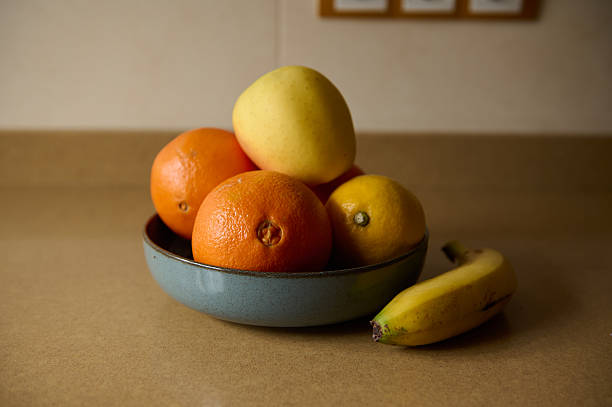 A still life of a blue ceramic bowl filled with citrus and apples, with a banana resting nearby. Warm kitchen lighting highlights fresh fruit for food, wellness, and home decor themes.