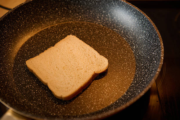 A single slice of white bread sits in a speckled frying pan, ready to toast. Warm oil surrounds it, suggesting a quick morning meal in a home kitchen.