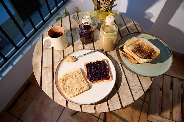 Sunny balcony breakfast scene with toast topped with jam and peanut butter, a mug of coffee, jars of spreads, and a wooden table for a warm, casual morning moment.