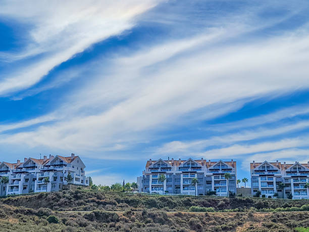 A row of contemporary white apartment buildings with terracotta roofs lines a steep coastal hillside, featuring multiple levels of balconies and windows overlooking a dry, shrub-covered landscape. The scene is set outdoors during a sunny daytime in summer, with a vibrant blue sky streaked by thin white clouds. No people are present, emphasizing the architectural design and serene environment. This image suits commercial uses in real estate promotions, travel brochures for Mediterranean-style destinations, architectural portfolios, or lifestyle content highlighting luxury coastal living and vacation properties.